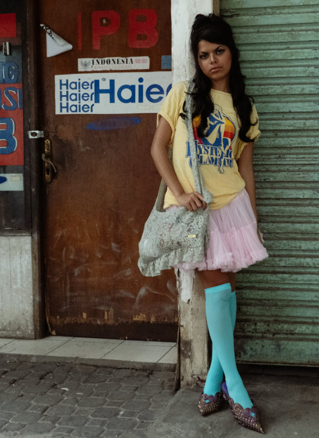 Woman standing against a door with Haier branding, wearing a yellow shirt and pink skirt with her lace mint green bag