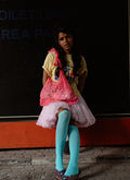 Person wearing pink lace bag in a yellow shirt, pink skirt, and blue tights sitting against a dark wall.