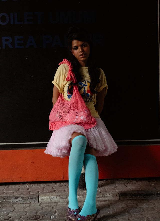 Person wearing pink lace bag in a yellow shirt, pink skirt, and blue tights sitting against a dark wall.